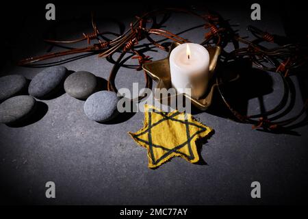 Holocaust-Gedächtnisfeiertag. Stacheldraht, Steine, gelber Stern und brennende Kerze auf schwarzem Hintergrund Stockfoto