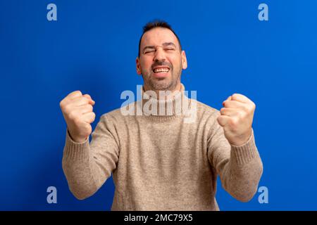Fröhlicher, aufgeregter Hispanic mit Rollkragenpullover, isoliert auf blauem Hintergrund stehend, um den Erfolg mit erhobenen Fäusten und geschlossenen Augen zu feiern Stockfoto