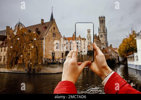 Eine Touristin fotografiert mit ihrem Handy historische Gebäude und einen Kanal in Brügge, Belgien. Stockfoto