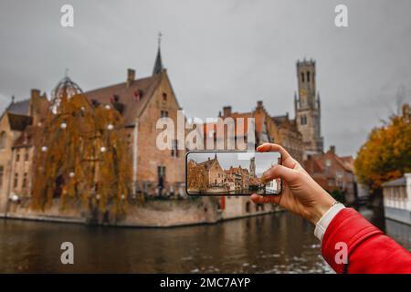 Eine Touristin fotografiert mit ihrem Handy historische Gebäude und einen Kanal in Brügge, Belgien. Stockfoto