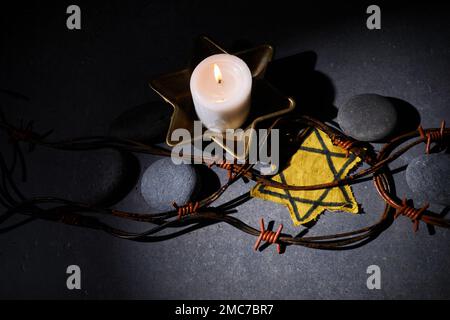 Holocaust-Gedächtnisfeiertag. Stacheldraht, Steine, gelber Stern und brennende Kerze auf schwarzem Hintergrund Stockfoto