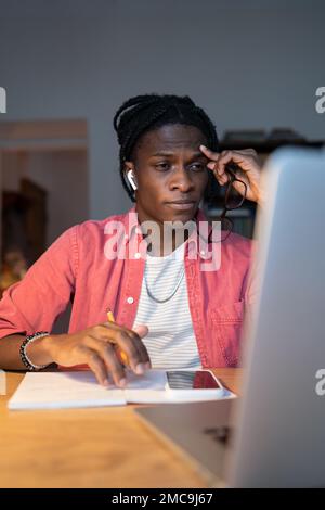 Ein müder, schwarzer, junger Mann, der auf die Stirn blickt und auf den Laptop-Bildschirm schaut, sitzt mit einem Bleistift am Tisch. Stockfoto