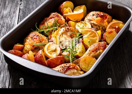 Zitronen-Knoblauch-Hühnchen mit gerösteten Kartoffeln, Gewürzen und Kräutern auf einer Blechpfanne auf einem dunklen Holztisch Stockfoto