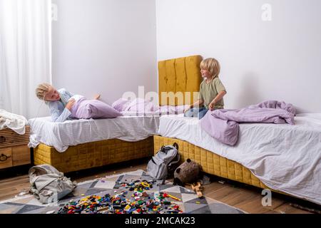 Geschwister, die Spaß haben, lachen, Junge und Mädchen spielen Kissenschlacht in einem dreckigen Kinderzimmer, Kinder spielen unter den vielen Spielzeugen zu Hause, viele Spielzeuge und Stockfoto