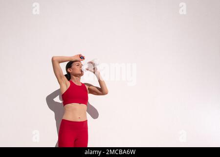 Sportliches weibliches Trinkwasser nach Bewegung und Laufen an einer weißen Wand Stockfoto