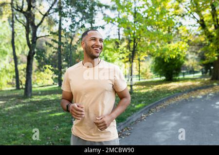 Ein fröhlicher und erfolgreicher hispanic, der im Park joggt, ein Mann, der an einem sonnigen Tag läuft, lächelt und glücklich ist, eine Outdoor-Aktivität zu haben. Stockfoto