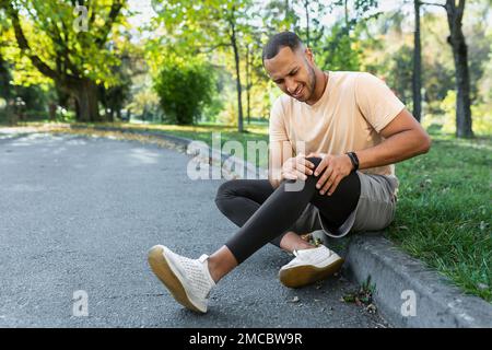 Ein Mann hat beim Joggen ein Bein verletzt, ein afroamerikanischer Mann sitzt auf dem Boden, massiert Muskelschmerzen mit den Händen, verärgert und verletzter Läufer. Stockfoto