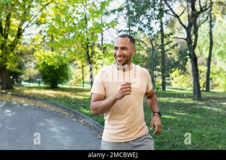 Ein fröhlicher und erfolgreicher hispanic, der im Park joggt, ein Mann, der an einem sonnigen Tag läuft, lächelt und glücklich ist, eine Outdoor-Aktivität zu haben. Stockfoto