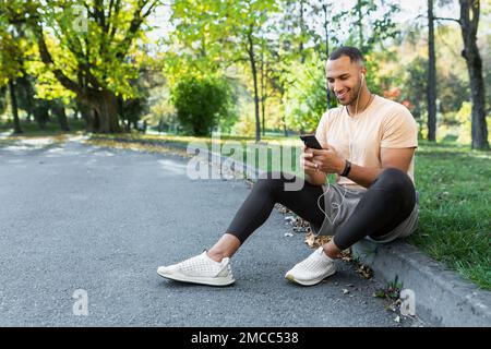 Glücklicher und zufriedener Mann nach Joggen und Fitness-Kurs sitzend und mit Smartphone, afroamerikanischer Mann mit Kopfhörern, der Online-Podcasts, Hörbücher und Musik hört, am Telefon tippt. Stockfoto