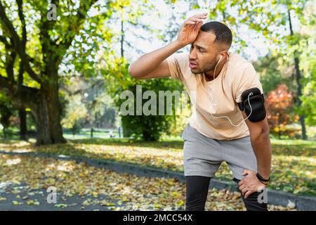 Müder Mann ruht sich nach Joggen und Fitness-Kurs im Park aus, hispanischer Mann, der telefoniert und Musik und Podcasts in Kopfhörern hört, und atmet müde und erschöpft. Stockfoto
