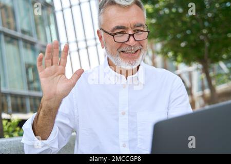 Lächelnder älterer Geschäftsmann, der auf ein Notebook schaut und bei einem Videoanruf mit der Hand winkt. Stockfoto