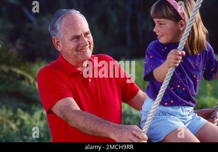 Opa drängt Mädchen, Enkelkind auf einer Schaukel, die sich beide lächeln sehen Stockfoto