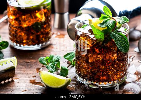 Rum-Cola-Cocktail mit starkem Alkohol und Eis, garniert mit Minze und Limette in Glas. Holzhintergrund, hartes Licht Stockfoto