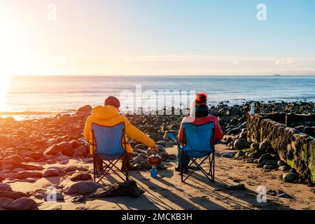 Zwei Leute auf Liegen zusammen am Winterstrand, sonniger blauer Himmelshorizont. Britischer kalter Winter. Konzept des lokalen Tourismus. Stockfoto