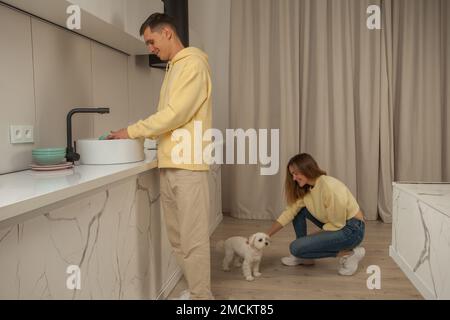 Glückliches, fröhliches Paar in der Küche in gelben Kapuzen, Mann, der das Geschirr wäscht und Frau, die sitzt und kleinen weißen Hund streichelt Stockfoto