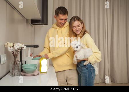 Portrait eines glücklichen Paares in gelben Kapuzen, Mann wäscht das Geschirr, Frau hält kleinen weißen Hund, Geschirrspülmittel mit leerem Etikett ist in der Nähe Stockfoto