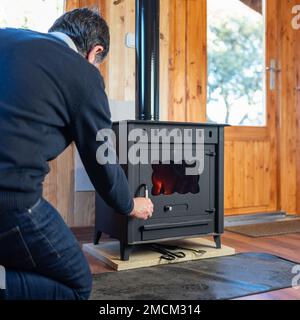Ein Mann öffnet die Tür eines rustikalen Gusseisenherds, um das Feuer zu entzünden. Stockfoto