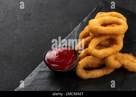 Calamari-Ringe frittiert in Panade. Knusprige Tintenfischringe auf dunklem Hintergrund mit Sauce. Speicherplatz kopieren. Stockfoto