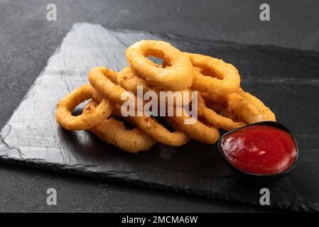 Calamari-Ringe frittiert in Panade. Knusprige Tintenfischringe auf dunklem Hintergrund mit Sauce. Stockfoto