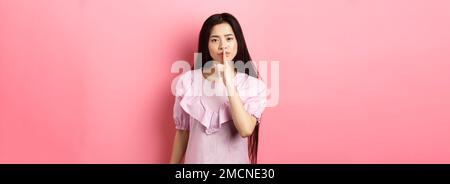 Cute asiatische Mädchen sagen, ruhig zu sein, Schelten laute Person, zeigt Shush Zeichen mit Finger auf die Lippen gedrückt, im Kleid auf rosa Hintergrund stehen Stockfoto