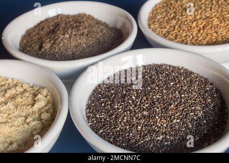 Nahaufnahme eines kleinen Tellers gefüllt mit Chiasamen, umgeben von Schüsseln aus ganzen und pulverförmigen Chiasamen und Leinsamen. Essen und gesunde Ernährung. Stockfoto