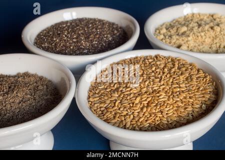 Nahaufnahme eines kleinen Tellers gefüllt mit Leinsamen, umgeben von Schüsseln aus ganzen und pulverförmigen Flachs- und Chiasamen. Essen und gesunde Ernährung. Stockfoto