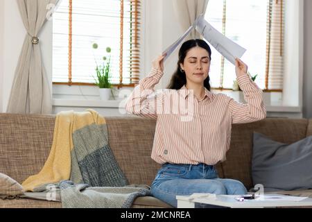 Porträt einer hübschen jungen Weißen, die auf dem Sofa sitzt und Dokumente in Form eines Dachs über ihren Kopf hält. Konzept von Kredit und Hypothek. Stockfoto