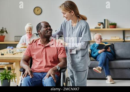 Ehrenamtlich mit einem afroamerikanischen Mann mit Behinderung während ihrer Arbeit im Pflegeheim im Raum sprechen Stockfoto