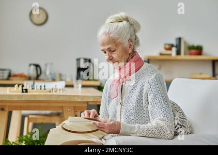 Ältere Frau, die sich auf ihr Handwerk konzentriert, sie sitzt auf einem Sessel und stickt Stockfoto