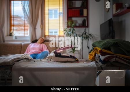 Heißes Bügeleisen auf dem Bügelbrett. Im unscharfen Hintergrund schläfende Frau auf dem Sofa. Konzept der Brandgefahr durch elektrische Haushaltsgeräte. Stockfoto