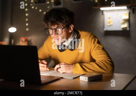 Attraktiver arabischer Student, der auf einem Laptop lernt, Notizen in einem Notizbuch schreibt und nachts im Schlafsaal sitzt. Speicherplatz kopieren Stockfoto