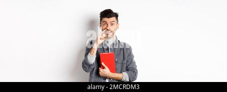 Junger Kerl Kommissionierung Nase und halten Buch, schauen dumm an der Kamera, stehen vor weißem Hintergrund Stockfoto