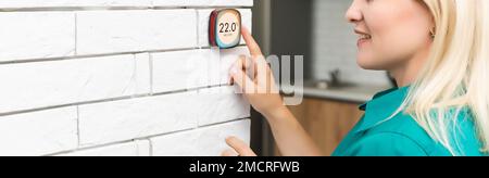 Frau, die die Heiztemperatur mit einem modernen, kabellosen Thermostat reguliert, der an der weißen Wand zu Hause installiert ist. Smart Home Heizung Regelkonzept Stockfoto