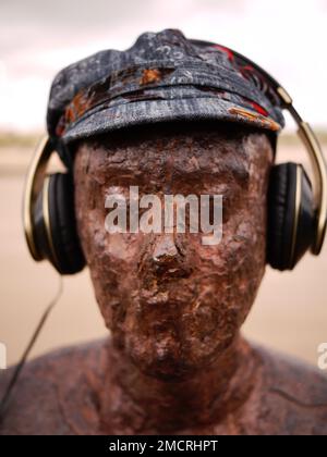 Eine von Antony Gormleys rostigen Eisenfiguren, Teil seiner Kunstinstallation an einem anderen Ort, trägt Hut und Kopfhörer. Crosby Beach, Merseyside Stockfoto