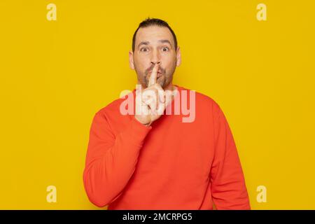 Ein gutaussehender Mann in einem roten Pulli, hält sich den Zeigefinger an die Lippen, versucht die Verschwörung zu halten, schaut zur Seite und sagt: "Sei still. Stockfoto