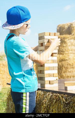Seitenansicht eines Jugendlichen Jungen, der Brettspiel Jenga spielt, der im Sommer an sonnigen Tagen einen Turm aus Holzblöcken im Freien in der Nähe von Heuhaufen auf dem Feld baut. Brettspiele Stockfoto