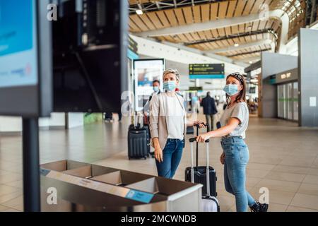 Zwei Reisende Frauen tragen Schutzmasken und diskutieren am Flughafen Faro über das Fluginformationsbrett Stockfoto