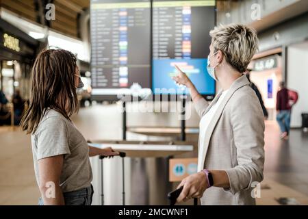 Zwei Reisende Frauen tragen Schutzmasken und diskutieren am Flughafen Faro über das Fluginformationsbrett Stockfoto