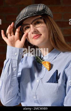 Hübsche, sehr junge Frau mit langen braunen Haaren trägt ein blaues Hemd mit Taschentuch und Schleife sowie eine karierte Mütze. Mit der rechten Hand Stockfoto