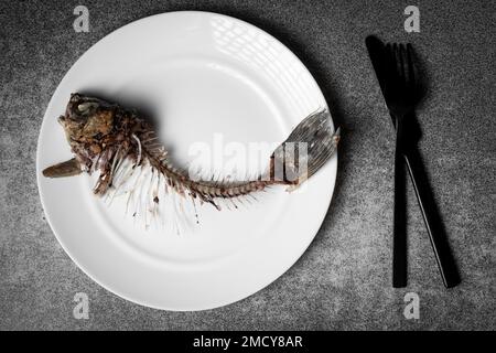 Gebratenes Fischskelett, Wirbelsäule und Knochen auf einem weißen Teller mit schwarzem Messer und Gabel auf grauem Hintergrund. Stockfoto