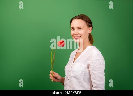 Eine hübsche Frau mittleren Alters, 40 Jahre alt, hat eine Blume - eine rote Tulpe auf grünem Hintergrund. Sie lächelt und schaut in die Kamera. Platz für dich Stockfoto