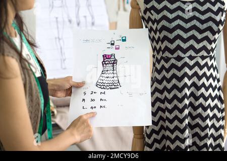 Junger Modebesitzer, der Bestellungen von einem Muster für Stoff überprüft, verglichen mit dem Kleid auf der Schaufensterpuppe. Stockfoto