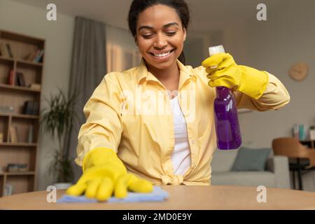 Fröhliche afroamerikanische Frau in Gummihandschuhen, Reinigungstisch im Wohnzimmer, mit Spray und Staubtuch Stockfoto