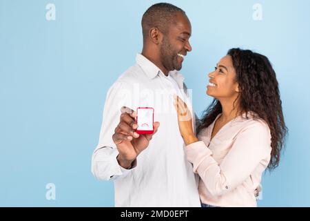 Liebevolles schwarzes Paar, das sich umarmt und anschaut, ein Mann mittleren Alters, der eine Box mit Verlobungsring hält Stockfoto