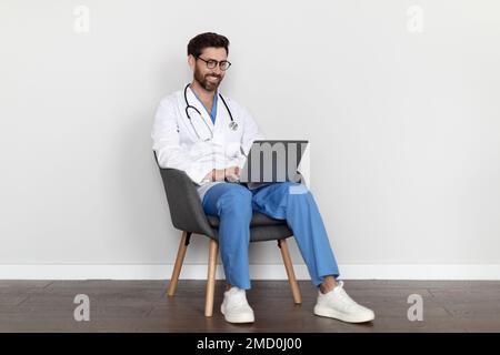 Telemedizin. Attraktiver Männlicher Arzt In Uniform, Sitzt Im Stuhl Und Benutzt Laptop Stockfoto