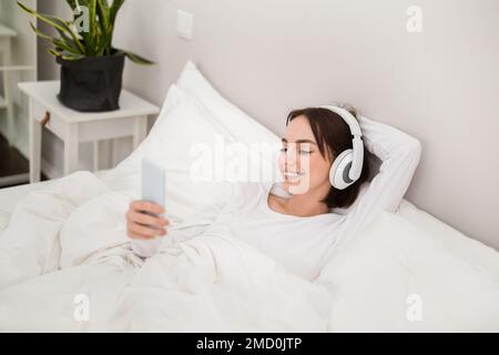 Glückliche junge Frau, die im Bett mit Telefon und Kopfhörern ruht Stockfoto