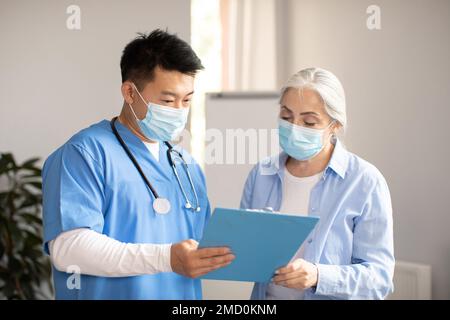 Ernster koreanischer Arzt mittleren Alters in Schutzmaske, der der alten weissen Patientin ein Vertragszeichen gibt Stockfoto