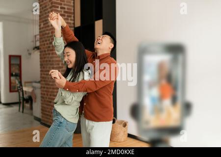 Verspielte asiatische Ehepartner übertragen oder zeichnen Inhalte auf, während sie vor der Smartphone-Kamera tanzen, Kopierbereich Stockfoto