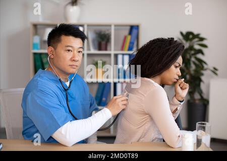 Ein seriöser, reifer koreanischer Arzt hört auf die Atmung von einer hustenden schwarzen Millennialdame in der Klinik Stockfoto