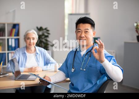 Glücklicher koreanischer Arzt mittleren Alters, der ein gutes Zeichen mit der Hand zeigt, und das Behandlungsergebnis einer alten europäischen Frau genehmigt Stockfoto